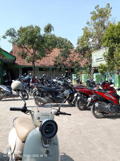 This image displays an outdoor motorcycle parking area located at a facility identified by a sign as "NEGERI 2 JAGAPURA KULON" in Jagapura Kulon, Indonesia. In the immediate foreground, a light cream-colored 'PACIFIC' brand retro-style scooter is partially visible, angled slightly left, showcasing its handlebars, headlight, and part of the seat. The midground is dominated by a dense arrangement of approximately two dozen parked motorcycles and scooters, primarily in black, dark red, and grey, resting on a light grey paved surface. In the background, a building with a green-painted lower facade and a reddish-brown tiled roof is partially visible, partially obscured by a large, leafy green tree. To the right, another section of a similar building and a green and white sign with Indonesian text are present amidst further green foliage. The scene is brightly illuminated by direct sunlight under a clear blue sky, casting distinct shadows. No individuals are visible within the frame.