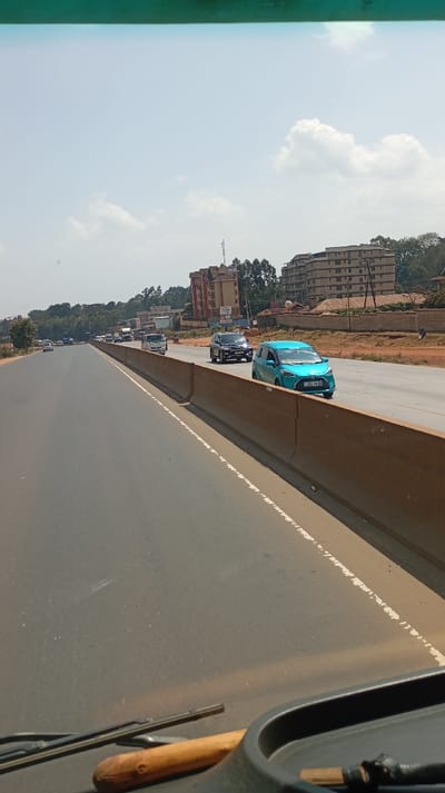 The image displays a multi-lane divided highway in Sigona ward, Kenya, viewed from inside a vehicle through its windshield. The top of the vehicle's dashboard, a windshield wiper, and a wooden object are visible in the foreground. The highway features asphalt lanes separated by a concrete barrier. On the right side of the barrier, a bright turquoise Toyota Sienta with Kenyan license plate KDJ 125R is visible, followed by a dark-colored SUV and a white van further in the distance. Traffic on this side moves generally in the same direction as the camera. On the left side of the barrier, multiple lanes of asphalt road recede, with light traffic present. The background features a raised dirt embankment and several multi-story buildings, including a reddish-brown structure and a lighter-colored building, interspersed with trees. The sky is partly cloudy with patches of clear blue. No individuals are distinctly visible.