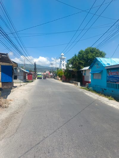 The image depicts a street scene in Tenggela, Indonesia, under a clear blue sky. A paved road runs through the center, lined with buildings on both sides. On the left, several small structures with corrugated metal roofs and simple facades are visible, one with a blue panel. On the right, a bright blue building with a white-tiled roof stands out, featuring a sign with the text "QACCO-R FISHING" and some Indonesian text below. Further down the road, a white tower-like structure with arches and a domed top is visible, possibly a landmark or a religious building. A green truck and a three-wheeled vehicle are on the road, along with several pedestrians. Overhead, a dense network of electrical wires crisscrosses the sky. The background shows a line of trees and rolling hills under a bright sky with scattered clouds. The overall scene suggests a warm, sunny day in a semi-urban or rural area.