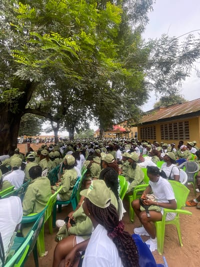 The image depicts a large outdoor gathering of young adults, many of whom are wearing identical light green uniforms and caps, suggesting a uniformed service or group. They are seated on bright green plastic chairs arranged in rows under a canopy of large trees. In the background, a yellow building with barred windows and a weathered roof is visible, along with other structures and more trees. The ground appears to be dirt or sand. Many individuals are facing forward, seemingly attentive to an event or presentation happening off-camera. The overall atmosphere suggests an organized event, possibly a training or orientation program, taking place in a communal or educational setting within Ise - Ekiti, Nigeria. The presence of the uniforms strongly indicates a uniformed youth service or similar organization prevalent in Nigeria.