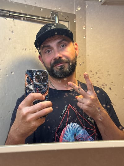 A man wearing a black baseball cap and a black t-shirt with a graphic of a nautilus shell and geometric lines is taking a selfie in a mirror. He has a beard and is holding a smartphone with a dragon-themed case in his left hand and making a peace sign with his right hand. The mirror is speckled with water droplets, and the background appears to be a bathroom with beige tiled walls and a metallic fixture above the mirror. This image is not located in Pevek, Russia.