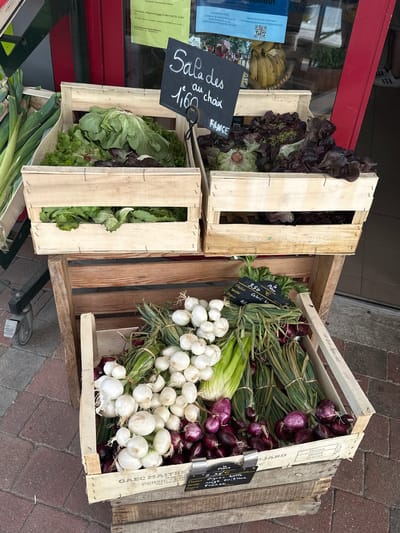 An outdoor market stall in Ouville-la-Rivière, France, displays fresh produce for sale. In the foreground, a wooden crate holds white onions and red onions, with bundles of green onions and celery also visible. Attached to the crate is a price tag indicating "Pièce 2.35 € Oignon rouge ou blanc" and "FRANCE". Above this, another wooden crate contains leeks.
Higher up, two wooden crates are stacked. The top left crate is filled with various types of lettuce. A black chalkboard sign placed on top of these crates reads "Salade au choix 1,60 €". The top right crate also contains a selection of leafy greens. The stall is set against the backdrop of a building with a red door, and a window displays posters. The ground is paved with reddish-brown bricks. A metal cart with wheels is partially visible on the left side of the image.
