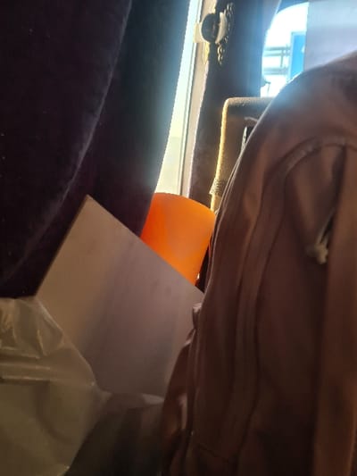A close-up view shows a tan backpack above a partially visible orange object. Below, a white package covered in plastic wrap sits on a dark purple, plush surface. Light enters from a window, illuminating part of the scene. The background shows glimpses of structures outside. The composition emphasizes texture and the layering of objects.