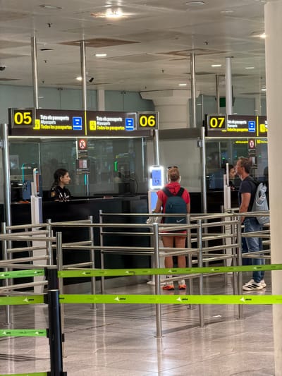This media file captures a scene at an airport's passport control area, likely at El Prat de Llobregat, Spain. The image is vertically oriented, showing multiple passport control lanes with automated gates and stanchions guiding a queue.

In the foreground, bright green "aena" branded barriers direct the flow of people. The main subjects are individuals in different lanes. In one lane, a man with short brown hair, wearing a dark t-shirt and blue jeans, is interacting with a passport scanner. Further down, a woman with blonde hair tied back, wearing a red t-shirt and denim shorts, is also at a scanner. In another lane, a woman with dark hair pulled back, wearing a dark uniform with patches that suggest she is a security or immigration officer, is looking upwards, possibly at a screen or a colleague.

Visible text includes "05", "06", and "07" on yellow signs, indicating the lane numbers. Below these numbers, text in Catalan and Spanish reads "Tots els passaports", "All passports", and "Todos los pasaportes". Blue rectangular signs with a passport icon are also present, along with a red circular sign with a diagonal line, indicating a prohibition.

The setting is indoors, with polished grey tiled flooring and modern airport infrastructure. The lighting appears to be artificial, typical of an airport terminal, suggesting it could be any time of day. The overall mood is one of transit and processing, with individuals focused on passing through immigration. The presence of automated gates and clear signage points to an efficient, organized system.