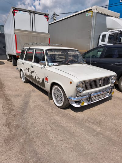 A white vintage Lada 2102 station wagon is parked outdoors on a rough, light-colored paved surface, likely a commercial lot in Molokovo, Russia. No people are visible in the scene.
The Lada is the central subject, customized with a black mountain and forest silhouette decal running along its side panels. A red logo featuring a sun and the Russian word "ЭКСПЕДИЦИЯ" (EXPEDITION) is present on its front fender. It features white-wall tires paired with silver multi-spoke rims and a chrome roof rack. Small yellow fog lights are mounted below the front bumper. A "05" sticker is visible on the windshield, along with a partially visible phone code "(495)". The car's Russian license plate reads "E274KE 550 RUS", indicating the Omsk Oblast region.
Behind the Lada, two large semi-trailers are parked. One is a white box trailer with a red trim and a sticker visible on its rear doors stating "ПРОДАЖА, СЕРВИС, TRADE-IN АВТОМОБИЛЕЙ ГАЗ" (Sales, Service, Trade-In of GAZ vehicles). The other is a grey curtain-side trailer with a "ГРЮНВАЛЬД ПРИЦЕПЫ" (GRUNWALD TRAILERS) sticker on its side, suggesting a trailer manufacturer. Part of a dark-colored SUV or crossover is visible to the right of the Lada.
The scene takes place during the daytime under a partly cloudy but bright sky, casting distinct shadows from the vehicles. The background shows parts of industrial or commercial buildings, including one with blue siding. The overall setting suggests a parking or service area for vehicles.