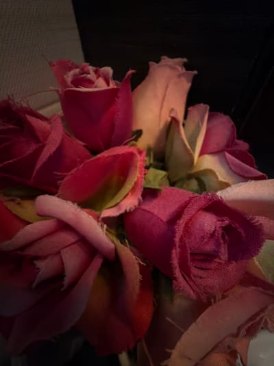 A cluster of fabric roses in varying shades of pink and blush fills the frame. The roses exhibit a deliberately frayed and vintage texture. Green leaves accent the blooms, adding a touch of contrast. A dark, possibly black, background looms on the left, while a neutral, textured surface sits beneath the roses. The lighting is subdued, emphasizing the depth and texture of the floral arrangement.