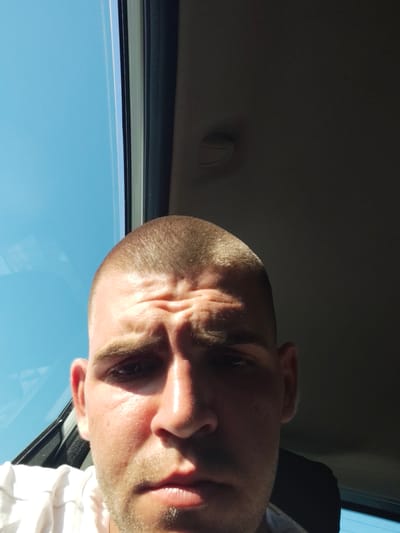 A close-up, eye-level selfie depicts a fair-skinned man with a buzz cut and a short beard. He is positioned centrally, with his face occupying most of the frame. The man's expression is neutral, and his eyebrows are furrowed, suggesting concentration or concern. He is wearing a white t-shirt. The background is divided into two distinct areas: on the left, a bright blue sky is visible through a window, and on the right, the dark interior of a vehicle, possibly a car, is apparent. A sun glare illuminates the left side of the man's face. The image was taken outdoors, likely inside a vehicle in Carrù, Italy.