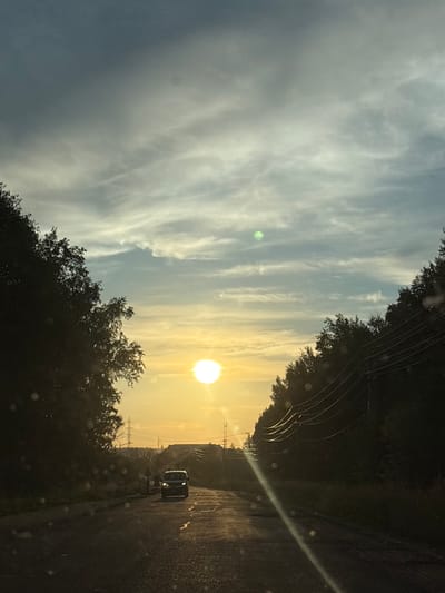 The image depicts a view from a vehicle driving on an asphalt road towards a bright sunset in Chigiri, Russia. The sun, a brilliant white orb, is low in the sky, casting a warm, golden light. Wispy, scattered clouds streak across the sky, with the upper portion of the sky appearing a darker, grayish-blue.
On either side of the road, dense trees, silhouetted by the setting sun, create a tunnel-like effect. On the right side, a series of power lines run parallel to the road, supported by poles. In the distance, a white minivan is visible on the road, its headlights illuminated. Further in the distance, there are signs of infrastructure, including what appear to be power line towers. There are also a few faint water droplets visible on the windshield.