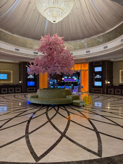 The image depicts an opulent indoor space, likely a casino or a high-end entertainment venue. Dominating the scene are luxurious design elements: a grand, multi-tiered chandelier hangs from a draped ceiling, while a large, artificial cherry blossom tree with pink flowers adds a touch of whimsical elegance. The floor is adorned with a striking geometric tile pattern in light beige and dark marble.
In the background, a section of the venue is visible with slot machines and illuminated screens displaying various graphics and text. A decorative, quilted seating area is positioned near the center, and several yellow "wet floor" signs are placed on the floor, suggesting recent cleaning or a potential hazard. The lighting is predominantly warm and artificial, creating a glamorous and inviting atmosphere.
Visible text includes what appears to be a phone number on one of the screens in the background, "1 249 777 24". Another screen displays a stylized logo, possibly for a casino game. The overall impression is one of a sophisticated and lively entertainment environment. The image is oriented vertically, as if taken from a high angle looking down, or the camera was held sideways. No people are clearly visible in the image.