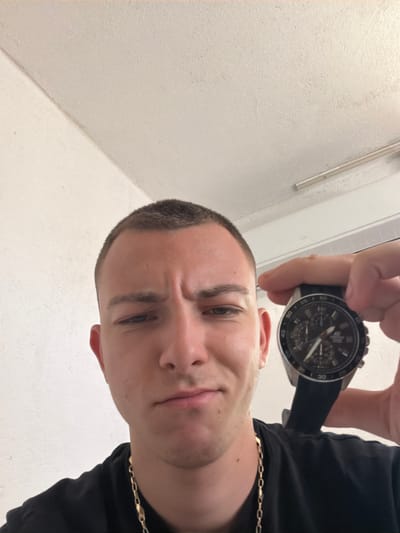 A low-angle selfie depicts a Caucasian male with short, dark hair, holding a black wristwatch up to the camera. He is wearing a black shirt and a gold chain. The background consists of a white wall with a molding. The location, Krivo Pole, Bulgaria, cannot be determined from this image.
