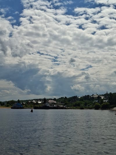 The image depicts a wide, dark body of water in the foreground, with gentle ripples on its surface. Along the far bank, a low-lying settlement is visible, characterized by a cluster of buildings with red and white roofs, interspersed with lush green trees. A prominent blue building stands out on the left side of the bank, near a sandy patch. Further to the right, several boats are docked along the waterfront. Above the settlement, a hilly landscape stretches into the distance, dotted with more trees and structures, including a church spire on the right side. The sky above is a dramatic expanse of dynamic, textured clouds, with patches of blue sky peeking through. The overall scene suggests a tranquil, possibly rural or small-town atmosphere on a summer day. The location is the city of Verkhny Uslon, Russia.