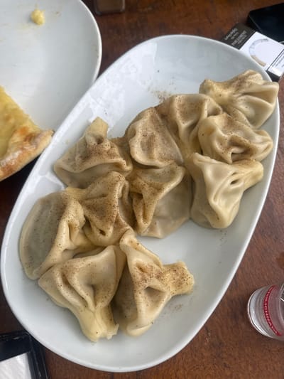 This media file showcases a close-up, overhead view of a white oval plate filled with Khinkali, a traditional Georgian dumpling. The dumplings are light-colored and appear to be sprinkled with black pepper. They are arranged in a cluster on the plate, with some overlapping.

In the background, a wooden table is visible, suggesting a dining setting. To the upper left of the plate, a partially visible cigarette pack is present, with the word "PARLIAMENT" legible on it. To the upper right, a small, clear shot glass is placed on the table, with red text that appears to read "STOLICHNAYA". The shot glass contains a clear liquid.

The overall scene suggests a casual meal, possibly at a restaurant or home, in Natakhtari, Georgia, given the context provided. The lighting suggests it could be daytime, but it's difficult to determine the exact time or weather conditions. The image focuses on the food, implying that eating is the primary activity. There are no people visible in the frame.