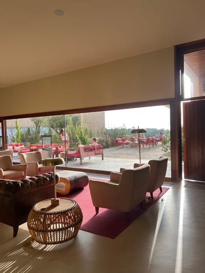 The image displays a modern lounge area within a building in Aljezur, Portugal, seamlessly opening onto an outdoor patio. The indoor space features a light grey concrete floor with a deep red rectangular rug. Furniture includes a dark brown leather tufted sofa on the left, an oblong wooden coffee table, and a rattan cylindrical side table supporting a brass-toned desk lamp. Two light brown upholstered armchairs are positioned with their backs to the right, facing away from the viewer. Strong natural light enters from a large window and a wooden-framed doorway on the right, casting distinct, elongated shadows across the floor. The ceiling is light beige and contains a single recessed light fixture.

Beyond a large, dark wood-framed glass partition, an outdoor patio extends, covered in light-colored gravel. This area is furnished with multiple sets of contemporary red lounge furniture, including sofas and individual chairs. At least five individuals are present on the patio. One female, dressed in light-colored clothing, stands in the mid-ground facing away from the viewer. Two males are seated on a red sofa, appearing to be in conversation. Two additional, less distinct figures are seated on red lounge furniture further in the background. Green foliage and trees are visible around the patio, and the light-colored facade of another building is present in the distant background. The scene suggests a leisure or hospitality setting bathed in bright daylight.