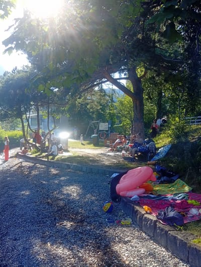 The image depicts a sunny day scene at a lakeshore, likely in Hergiswil (NW), Switzerland, given the context. The foreground is dominated by a pebbled beach with scattered debris and shadows from overhead trees. A stone embankment runs along the edge of the beach, on top of which several colorful blankets, towels, and inflatable toys are spread out. Among the inflatables is a large pink ring float and an orange float. A striped beach ball and a pair of goggles are also visible on the pebbles.

Moving further into the scene, a group of people are gathered under the shade of large trees. Some are seated on chairs and blankets, appearing to be engaged in conversation or relaxation. One individual, a woman with blonde hair, stands near the water's edge on the left, wearing a white top and an orange skirt. Further back, a group of people are seated around a table and some are standing. In the background, there's a glimpse of buildings, possibly houses or facilities, and more lush greenery. A playground structure with a swing set is visible to the right of the central gathering area, and a signpost with multiple information boards stands nearby. The overall atmosphere suggests a casual outing by the lake, possibly a picnic or a day at the beach. The bright sunlight creates strong highlights and shadows throughout the scene, indicating a clear and warm day.