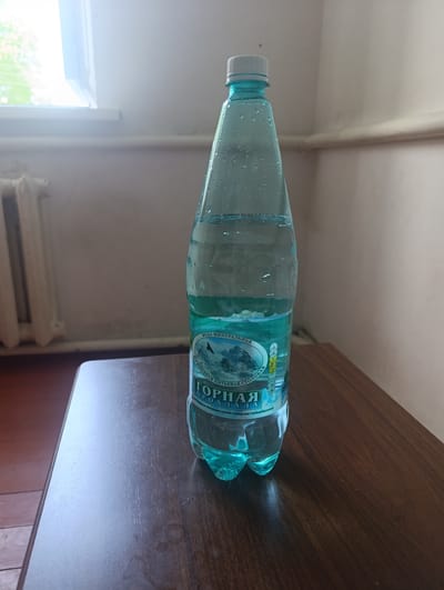 A full shot shows a plastic bottle of mineral water, "Gornaya Prohladnaya," standing on a wooden table. The bottle is approximately two-thirds full and has condensation on its surface. The label features a picture of mountains and text in Cyrillic. The bottle is clear blue and has a white cap. The table is dark brown and has a wood grain pattern. In the background, a white radiator and a window with bright sunlight are visible. The setting appears to be an indoor space, possibly a room in Gizel, Russia, suggested by the presence of Russian text on the bottle. There are no people or animals in the image.