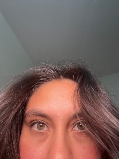 The image is a close-up, low-angle shot of a person's eyes and forehead, with their dark hair framing their face. The background is a plain, greyish wall that appears to be a ceiling or an upper wall, with a subtle shadow indicating a corner. There is no discernible information about the location within San Luis, Peru, as the image focuses solely on the individual's facial features and hair against a neutral background.