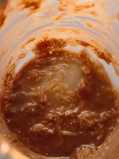 The image is a close-up, top-down view inside a clear plastic container, likely a cup or jar. The interior of the container is smeared with a reddish-brown substance, possibly food residue, mixed with a lighter, creamy liquid. The texture suggests some particulate matter, such as grains or small fibers, are suspended within the mixture and also cling to the sides of the container. The lighting creates reflections and highlights on the slick surfaces. There is no discernible geographical location or context for Dzerzhinsk, Russia within the image itself.