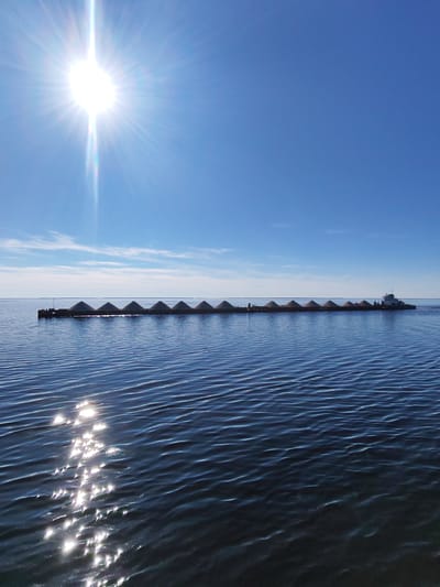 The image depicts a vast expanse of water under a clear blue sky in the city of Gorlovo, Russia. An intensely bright sun, positioned in the upper left quadrant, emits strong light rays and lens flare, creating shimmering reflections on the water surface in the lower-left foreground. A long train of approximately twelve dark-hulled barges, each carrying a symmetrical, light-colored conical mound of bulk material, traverses the midground horizontally. A white and dark-hulled tugboat is positioned at the rear of the barge train, actively propelling the convoy. The water exhibits subtle ripples and waves, most pronounced in the areas reflecting sunlight. A flat horizon line defines the distant edge of the water, beneath a sky featuring scattered, wispy clouds along the left horizon. No people are discernible.