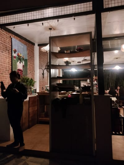 The image shows the interior of a restaurant at night, possibly in Kidapawan City, Philippines. In the foreground on the left, a woman in a dark top and pants is standing, looking at her phone. Behind her, a brick wall is decorated with a colorful tapestry depicting three figures carrying baskets of fruit. To the right of the woman, a tall, open shelving unit is filled with various items. The background reveals more of the restaurant, with tables, chairs, and other patrons visible, including a group of women seated at a table. Bamboo stalks can be seen in the far background. The lighting is dim, with overhead lights casting a warm glow.