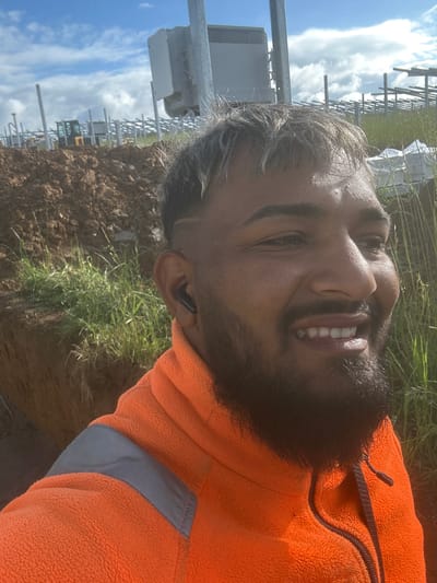 The image is a bright, outdoor selfie-style portrait of a man, taken during the day under a blue sky with white clouds. The man, smiling widely and revealing his teeth, appears to be of South Asian or Middle Eastern descent, with a dark beard and dark hair showing some graying at the temples. He is wearing a vivid orange fleece jacket, featuring a black zipper and a gray reflective strip on the lower part. A black earbud is visible in his right ear.
The background suggests an active industrial or construction site. In the foreground and midground, there is loose, brown earth, indicative of excavation, along with patches of green grass. Behind him, numerous metal structures, resembling frames, pylons, or scaffolding, extend into the distance. A white box-like electrical or communication unit is mounted on a pole to the left. Further back, a piece of yellow industrial machinery can be seen among the structures. The overall scene conveys a cheerful moment during outdoor work, likely at a construction or large-scale installation site in Weissach im Tal, Germany. There is no visible text in the image.
