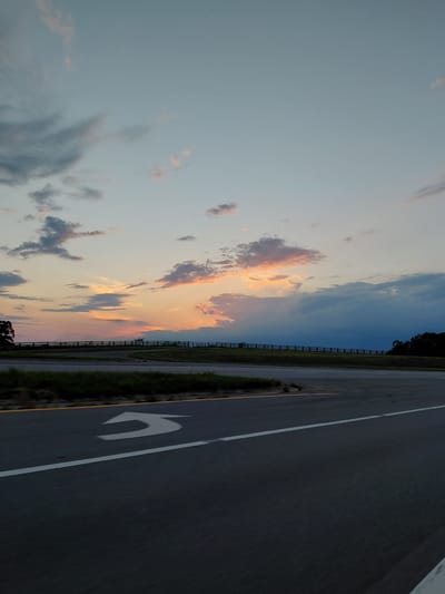 This image captures a sunset scene on a road in Lancaster, United States. The sky is a canvas of soft blues and grays, with streaks of pink and orange from the setting sun illuminating scattered clouds. A dark, horizontal fence stretches across the middle ground, with a line of trees silhouetted against the horizon. In the foreground, the asphalt road is visible, marked with white and yellow lines, and a white arrow indicating a turn is painted on the surface. The overall impression is one of serene twilight over a rural or semi-rural landscape.