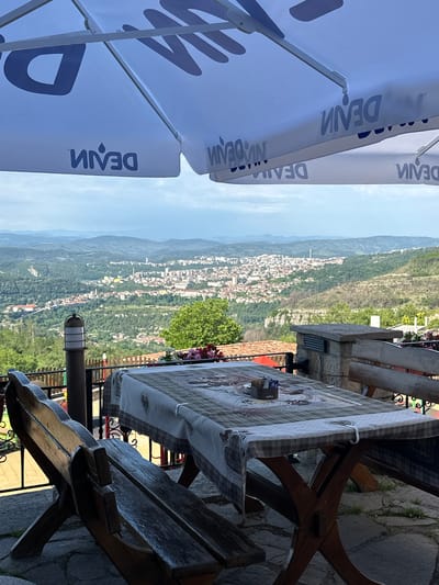 The image shows a scenic view of the city of Arbanassi, Bulgaria, from a vantage point on a terrace. In the foreground, a wooden picnic table is set with a checkered tablecloth, a salt and pepper shaker, and a small box of sugar packets. Two wooden benches are positioned on either side of the table. A large white umbrella, emblazoned with the blue "DEVIN" logo, provides shade for the seating area. The umbrella also features a repeating pattern of the "DEVIN" logo. Beyond the terrace, a vast cityscape unfolds, nestled within rolling green hills and mountains under a cloudy sky. The city appears to be densely populated with buildings of varying colors and architectural styles. A prominent tower-like structure is visible in the distance. The overall impression is of a serene outdoor dining or relaxation spot offering a panoramic view of the historic Bulgarian town.