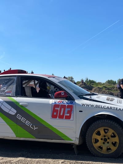 The image shows a white Geely rally car, number 603, at a rally event. The car has the inscription "GEELY" on the side and "www.BELCARTEAM.com" on the front. A driver wearing a red helmet is visible inside. The car appears to be on a dirt or gravel surface. There is a crowd of spectators in the background and blue sky. The Netherlands flag is visible on the window. The photo was taken in the Kanjiza Municipality, Serbia.