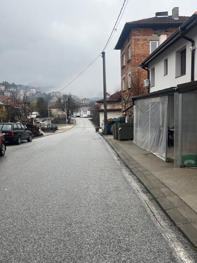 The image depicts a street scene in Srednogortsi, Bulgaria. The view is looking downhill on a wet, grey day, likely after or during rain. A two-story building with a brick facade and a white upper level is on the left side of the street. The lower part of the building appears to be undergoing renovation or construction. A covered outdoor storage area abuts the building. Cars are parked along the right side of the street, facing downhill. Utility poles and wires run alongside the street. In the distance, there is a hillside covered with buildings and trees, partially obscured by mist or fog.