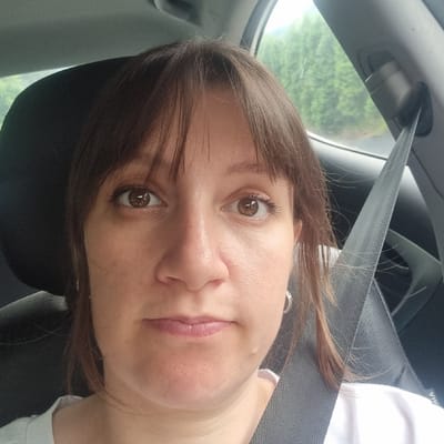 A medium shot captures a woman from the chest up, seated in the passenger seat of a car. She has shoulder-length, brown hair with bangs and is wearing a white t-shirt. Her eyes are brown and she is looking directly at the camera. A gray seatbelt is visible across her chest. The background shows green trees through the car window, suggesting a rural or suburban setting. The image appears to be taken in Gangnim-myeon, South Korea.