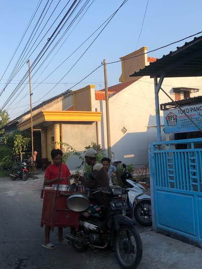 In Kalidawir, Indonesia, two young males and an older male are engaged in a transaction near a street. A young male, wearing a red t-shirt and light-colored slippers, stands to the left, appearing to be the vendor, with a food cart attached to a motorcycle. The cart, a red wooden structure, holds metal containers, one of which is covered by a lid. An older male, in a brown and tan shirt and a light-colored cap, is on the right, reaching towards the cart, possibly making a purchase or receiving food. Another young male in a light-colored shirt is partially visible behind the older male, observing the interaction. Two motorcycles are parked nearby: a black motorcycle in the foreground with a visible license plate "W 4499 PZ 11-17", and a white motorcycle behind it. To the right, a bright blue metal gate is prominent, and a shop sign above it reads "TOKO PANCA" and "BFS BOSS UNA FISHING STORE". The background features houses with orange tiled roofs and yellow decorative elements, and overhead, a tangle of electrical wires stretches across the clear blue sky. Trees and foliage are visible on the left side of the scene. The overall atmosphere suggests a typical street scene during daylight hours.