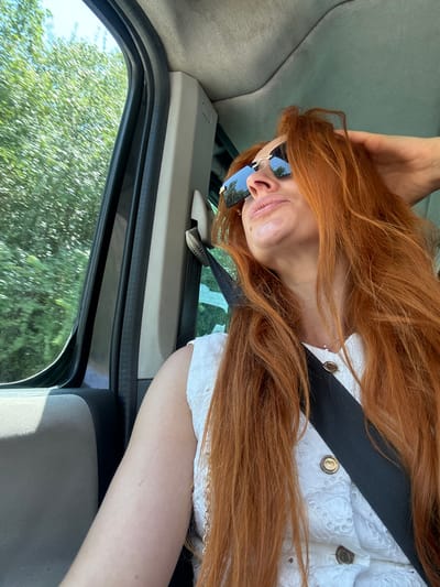 The image shows a woman with long, red hair sitting in a car. She is wearing a white, textured top with a black collar and gold buttons, and a pair of sunglasses with blue mirrored lenses. Her arm is resting on the car's interior. Through the open car window, lush green trees and a bright sky are visible, suggesting a sunny day. The scene appears to be a casual moment, possibly during a car journey. The interior of the car has a light gray fabric seat and a dark gray trim around the window. The angle of the shot, taken from inside the car and looking outwards, is unusual, with the woman's head tilted to the side. There is no visible text in the image. The location is Gornji Katun, Montenegro.