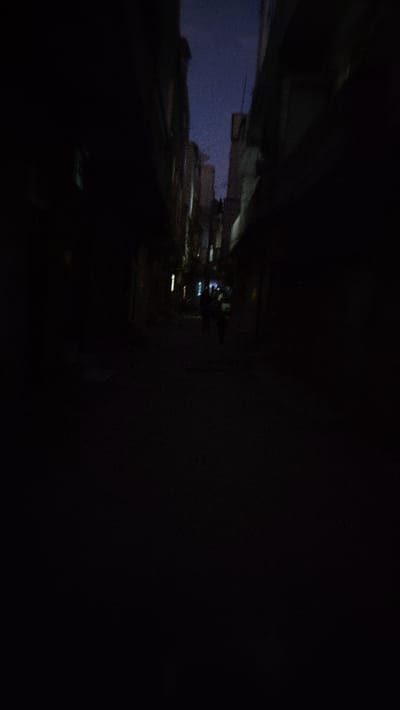 A dimly lit, narrow alleyway stretches into the distance, framed by buildings on either side. A faint blue glow hints at the evening sky above. Two blurred figures can be seen further down the alley, seemingly walking away. The foreground is dominated by darkness, obscuring details on the ground. A solitary light source illuminates a small section of the buildings, creating a sense of depth.