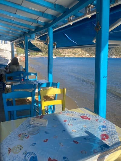 The image depicts an outdoor, covered dining area of a waterfront establishment located directly on a sandy beach in Απολλωνία, Greece. The structure is constructed with bright blue painted wooden beams and columns supporting a light-colored roof and blue retractable awnings.
In the foreground, a table covered with a light blue tablecloth adorned with red and blue fish patterns is visible. On this table, a small, light-colored rectangular block with the word "Reserved" inscribed in cursive is placed next to a clear glass ashtray. Beyond this table, several other tables with clear coverings and predominantly blue-painted wooden chairs are arranged. One chair is distinctly painted yellow.
One individual, seen from behind, with dark hair, is seated at a table further back within the dining area, wearing a light-colored top.
Beyond the dining area, a sandy beach extends into the clear, calm blue sea, where gentle waves are visible. Several small boats are moored in the water in the middle distance. In the far background, verdant hills rise under a clear sky, with sparse white buildings dotting the coastline along the base of the hills. Shadows cast by the blue structural elements indicate strong sunlight.