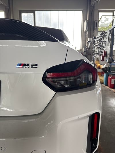 This is a close-up, vertically oriented, eye-level shot of the rear of a white BMW M2 car, possibly in a workshop or garage in Wettstetten, Germany. The car's rear light cluster is prominently featured on the left side of the frame. This cluster is sleek and modern, with a distinctive C-shaped red accent and a dark interior. Below the taillight, the "M2" badge is visible, with the "M" being a stylized emblem and the "2" incorporating the signature BMW M stripes in blue, red, and a lighter blue. To the left of the badge, a portion of a carbon fiber rear spoiler is visible, hinting at the car's performance focus.
The background is slightly out of focus, showing a workshop environment. To the far left, a window with multiple panes allows natural light to enter, illuminating shelves or racks filled with various automotive tools and equipment, including red and yellow canisters. The walls appear to be concrete or painted. The overall lighting suggests it is daytime, possibly late morning or afternoon, given the quality of the natural light filtering through the window. The car's surface shows some dust or water spots, indicating it might have been recently washed or is in a dusty environment. There is no direct indication of people or an ongoing event, suggesting a static display or preparation phase. The scene evokes a sense of automotive detail and craftsmanship.