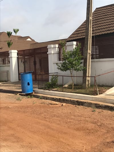 The scene is in Oko Erin, Nigeria, displaying a partially obstructed view of a two-story residential building. The building has a brown tiled roof, white support pillars, and brown slat-style fencing. A blue plastic barrel with a hole cut out of it sits in front of the building near a drain. A power line pole is situated close to the house. A small tree is present inside a fenced-off area. The ground is dirt, likely a road or driveway.