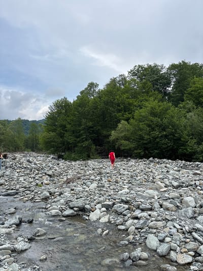 The image depicts a wide, rocky riverbed in Lagodekhi, Georgia, under an overcast sky. A person dressed in a red t-shirt and red shorts is observed walking barefoot across the expansive riverbed, moving away from the viewer towards a dense line of green forest. Another individual, partially visible on the far left, shows a torso in blue jeans and a light top, with their back to the camera. The riverbed is densely covered with an array of smooth, grey and white stones of varying sizes. A shallow, clear stream meanders through the foreground, reflecting the cloudy sky. Beyond the immediate forest, forested mountains are visible in the distance, partially obscured by low-lying clouds.