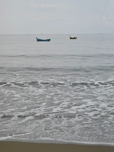 In Curehtunong, Indonesia, two small fishing boats are moored on a calm, grey sea under an overcast sky. The boat on the left is a bright blue, narrow vessel, while the boat on the right is dark green with yellow accents. The water is mostly still, with gentle ripples on the surface. Closer to the shore, the water breaks into white foam as it washes onto a sandy beach. The overall atmosphere is serene and quiet.