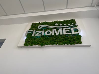 The image shows a wall-mounted sign with the text "FizioMED" displayed in prominent, silver 3D lettering. The lettering is placed over a backdrop of lush green preserved moss. Above the text, a stylized logo consists of a white wavy line with several circular dots. The sign is mounted on a white rectangular backing board. The setting appears to be an interior space, possibly an office or clinic, given the white walls and the ceiling with acoustic tiles visible at the top of the image. The lighting suggests it is daytime. No people are visible in the image. The location context is Suceava, Romania.