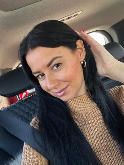 The image depicts an adult female positioned inside a vehicle, observed from the chest up. She possesses long, dark, straight hair, brown eyes, and is wearing a light brown, ribbed knit sweater. Her left hand, adorned with red nail polish, is raised toward her head, partially concealed by her hair near her right ear. Visible earrings include a small stud and a larger, gold-toned C-shaped earring in her left earlobe. A black seatbelt is diagonally positioned across her torso. The vehicle's interior features black leather seating with a quilted texture, a tan headliner with an overhead console, and a discernible red object in the distant background to the left. The subject maintains direct eye contact with a slight smile. The image contains no visual data to identify a specific location within Cham, Germany.
