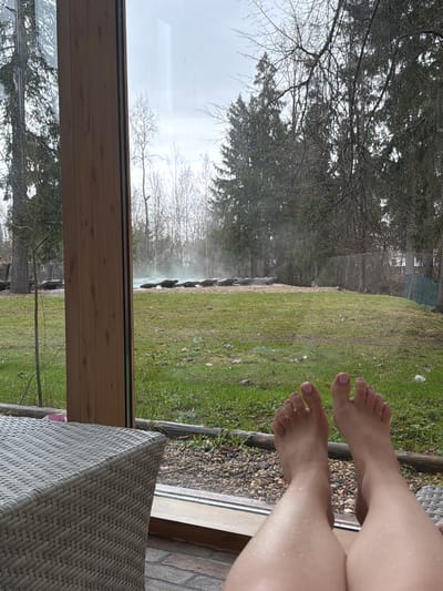 The image shows a partial view of a person's legs and feet, resting on a wicker chair or stool. The background consists of a view through a window, depicting a lawn area with green grass, trees, and possibly a body of water or a feature emitting steam or mist. The environment suggests a rural or suburban setting. The location is specified as Dulepovo, Russia, but there are no immediately discernible features in the image that confirm this geographical location. The person is likely relaxing or enjoying the view. The scene gives the impression of relaxation and leisure.