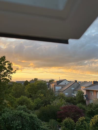 The image depicts a suburban landscape at dusk in Mondercange, Luxembourg. A substantial overhang, likely part of a building's structure, occupies the foreground, casting a shadow over the upper portion of the scene. Below this, a panorama of residential rooftops and lush greenery unfolds. The sky is a canvas of dramatic clouds, illuminated by the warm, golden hues of the setting sun, with patches of blue visible. The architecture appears to be modern, with pitched roofs and various building materials. Dense trees and gardens fill the spaces between the houses, suggesting a well-developed residential area with significant green spaces. No individuals or overt actions are captured within the frame.