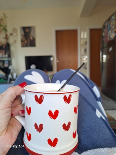 The image depicts an indoor residential scene from a first-person perspective. In the immediate foreground, a left hand with red manicured fingernails holds a white ceramic mug decorated with multiple red hearts and a red rim and base. The mug contains a light-colored, foamy beverage into which a slender black stirring stick is inserted. Below the mug, a person's upper legs are visible, covered by blue fabric loungewear featuring white heart patterns. A "Galaxy A54 5G" watermark is present in the bottom-left corner. The background is out of focus, revealing elements of a room including a dark television screen to the left, light-colored walls, two dark brown doors, and a partial view of a dark refrigerator on the right side. Blurred wall decorations, including a framed picture and a plant, are also discernible. The environment is an indoor living space. No specific identifying features are present to pinpoint a precise location within Xylofagou, Cyprus.