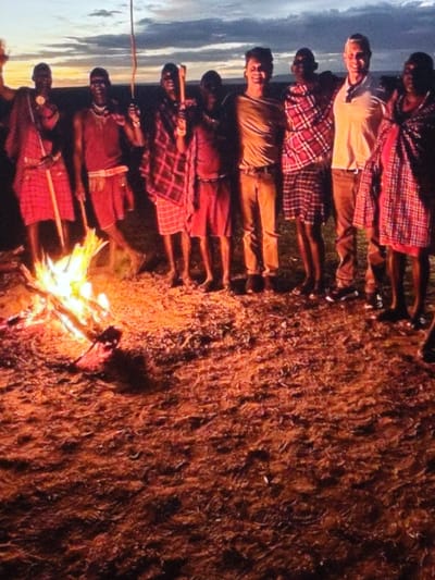 A group of nine individuals is gathered outdoors around a bonfire at dusk or night in the city of Dodoma, Tanzania. The scene is illuminated by the bright yellow and orange flames of the bonfire, positioned in the lower-left foreground. The immediate ground consists of dry earth and sparse, reddish-brown vegetation, glowing from the fire's light. The background reveals a twilight sky with bands of blue, grey, and faint orange clouds above a distant horizon.
Six individuals are adult males of dark complexion, dressed in traditional Maasai attire: red shukas, some solid red and others red and black plaid. Several wear beaded necklaces, and at least three are holding slender wooden staffs or clubs. The individual farthest to the left has one arm raised. Two other individuals, appearing to be non-African adult males, are present. One wears a light-colored t-shirt and dark trousers with a belt; the other wears a light-colored polo shirt and light-colored trousers. Both non-African males have their arms around the shoulders of adjacent Maasai individuals. All visible faces are smiling and directed towards the camera. The individuals are arranged in a horizontal line behind the bonfire, with one Maasai individual partially visible at the far right.