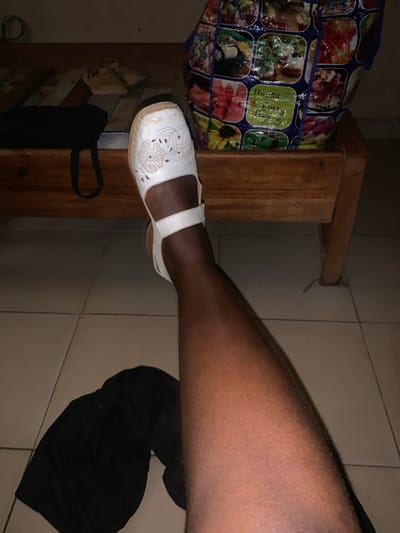 A dark-skinned human leg, identified as the right leg, extends diagonally upward from the lower left toward the upper right of the frame, viewed from a first-person perspective. The foot is encased in a white, closed-toe sandal with decorative cut-out patterns on the vamp and a secured instep strap. The sandal's toe area shows signs of wear and minor soiling. The leg exhibits visible hair. The immediate ground surface is a tiled floor with light-colored, square tiles. A crumpled piece of black fabric, consistent with clothing, rests on the tiled floor to the lower left of the leg. In the background, a light-colored wooden structure, resembling a bed frame or low bench, runs horizontally. On this wooden structure, a black bag with a strap is positioned to the left. Further to the right on the wooden structure, a pair of light-colored slip-on shoes are visible. A large, multicolored woven plastic bag, decorated with various images including parrots, flowers, and text that appears to include "Happy Every Day!!!", is situated on the wooden structure to the far right. The scene is dimly lit. This event is observed within the city of Zonkwa, Nigeria.