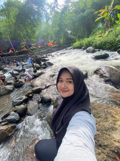 A woman in a black hijab smiles at the camera, positioned near a rocky stream. Flowing water rushes around her, reflecting light. A camping area with tents is visible in the background, surrounded by lush green trees. People are gathered near the stream, some appearing to play in the water. The camera angle suggests the photo may be a selfie.