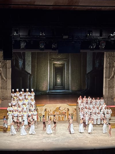 The image depicts a staged performance, likely a theatrical production, with an ancient Egyptian theme. The backdrop features a visual tunnel effect, suggesting a long corridor or temple interior, complete with hieroglyphic designs. The stage is populated by performers; one group is in the upper right of the shot, clad in white robes and headgear, while another grouping in the lower right wears similar costumes. Interspersed within these groups are performers in darker clothing. It appears the scene is lit with theater lights, visible on the left of the frame. This theatrical stage is located in San Borja, Peru.