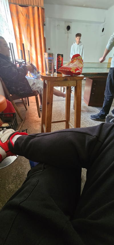 The image shows the interior of a building, likely a room with a table. Several individuals are present, including one in a white hoodie and another wearing black sweatpants with red and white shoes. The location is speculated to be Sveti Konstantin, Bulgaria. A wooden table is visible, with objects such as a bag of chips, a glass, and a can on it. Underneath the table, there is a chair with a person seated. The room appears to be naturally lit, with orange curtains or a covering visible. The flooring is concrete. Overall, it suggests a casual setting.