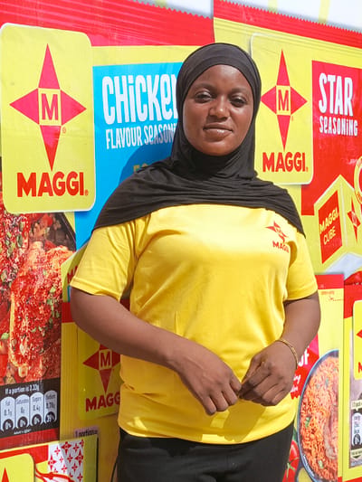 A woman is pictured standing outdoors, likely at a promotional event or a market stall in Kawaji, Nigeria, given the bright daylight and the commercial backdrop. She has dark skin and is wearing a black hijab that covers her head and neck, and a bright yellow short-sleeved t-shirt. The yellow t-shirt features a red Maggi "M" star logo on the left chest. Her hands are clasped in front of her, and she wears a thin gold-colored bracelet on her right wrist. She looks directly at the camera with a neutral expression.
The background is dominated by a large, colorful display featuring Maggi brand products. The display mimics large packets of Maggi seasoning, primarily in yellow, red, and blue colors. Prominently visible are the "MAGGI" brand name and its distinctive "M" star logo. Text on the display includes "CHICKEN FLAVOUR SEASONING" on a blue panel, and "STAR SEASONING" on a red panel. Another section shows "MAGGI CUBE."
Detailed nutritional information is visible on a dark panel, stating: "If a portion is 3.33g," followed by values for "Fat 0.1g 0%," "Saturates 0g 0%," "Sugars 0.4g 0%," and "Sodium 0.78g 39%," with "Adult's GDA" indicated. Other partial text includes "1 portion=", "OPEN HERE," and "Energy 25kJ 6kcal." Images of food, such as what appears to be jollof rice and a chicken dish, are also part of the vibrant product packaging illustrations on the display.