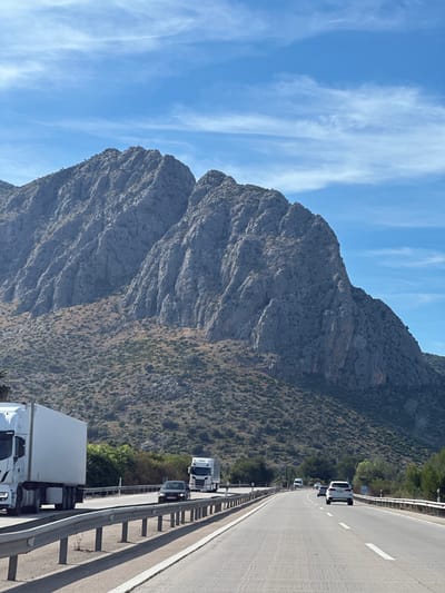 The image depicts a highway scene in Ondara, Spain, with a large mountain dominating the left side. The sky is blue with scattered clouds. A multi-lane highway runs on the right side, bordered by a metal guardrail. Several vehicles, including trucks and cars, are visible on the road. The mountain is rocky and partially covered with vegetation.