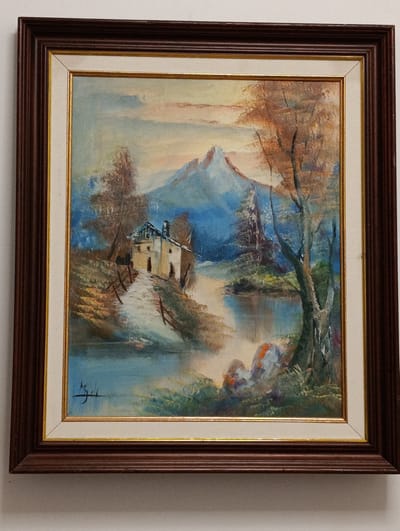 A framed oil painting on canvas depicts a serene landscape. In the foreground, a body of water reflects the sky and surrounding foliage. A pathway, possibly made of stone or gravel, leads uphill towards a small, light-colored house with a visible chimney. The house is nestled on a gentle slope, with sparse trees and fences lining the path. Further in the background, a majestic, snow-capped mountain rises against a hazy sky. The overall impression is one of natural beauty and tranquility. The painting is signed in black ink in the lower-left corner with what appears to be a stylized signature. The frame is dark wood with a ribbed texture. The painting is hanging on a plain white wall.